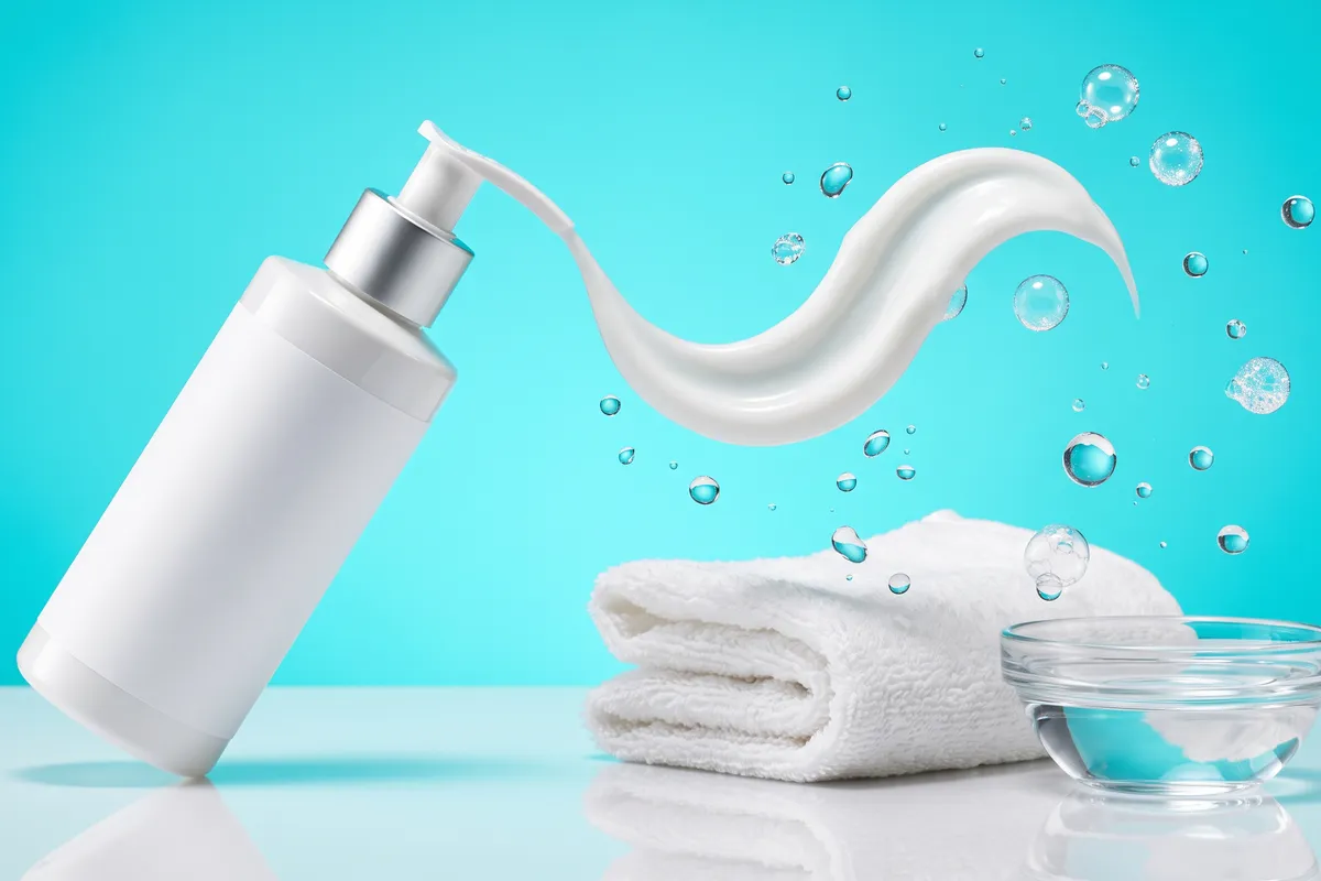 A ribbon of white cream cleanser curling out of an unbranded pump bottle beside a soft folded washcloth and clear water droplets on a bright aqua backdrop