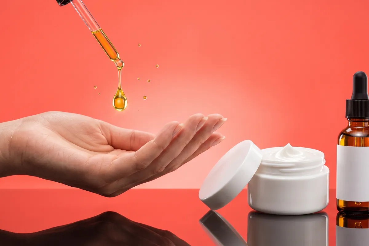 A single glossy drop of golden face oil falling from a glass dropper onto the fingertips of an open hand, beside an unbranded cream jar and amber bottle on a bright coral backdrop