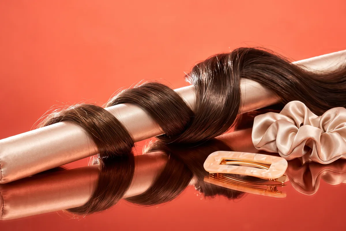 A satin heatless curling rod wrapped with soft brown waves beside two scrunchies and a pastel hair clip on a glossy surface against a bright coral backdrop