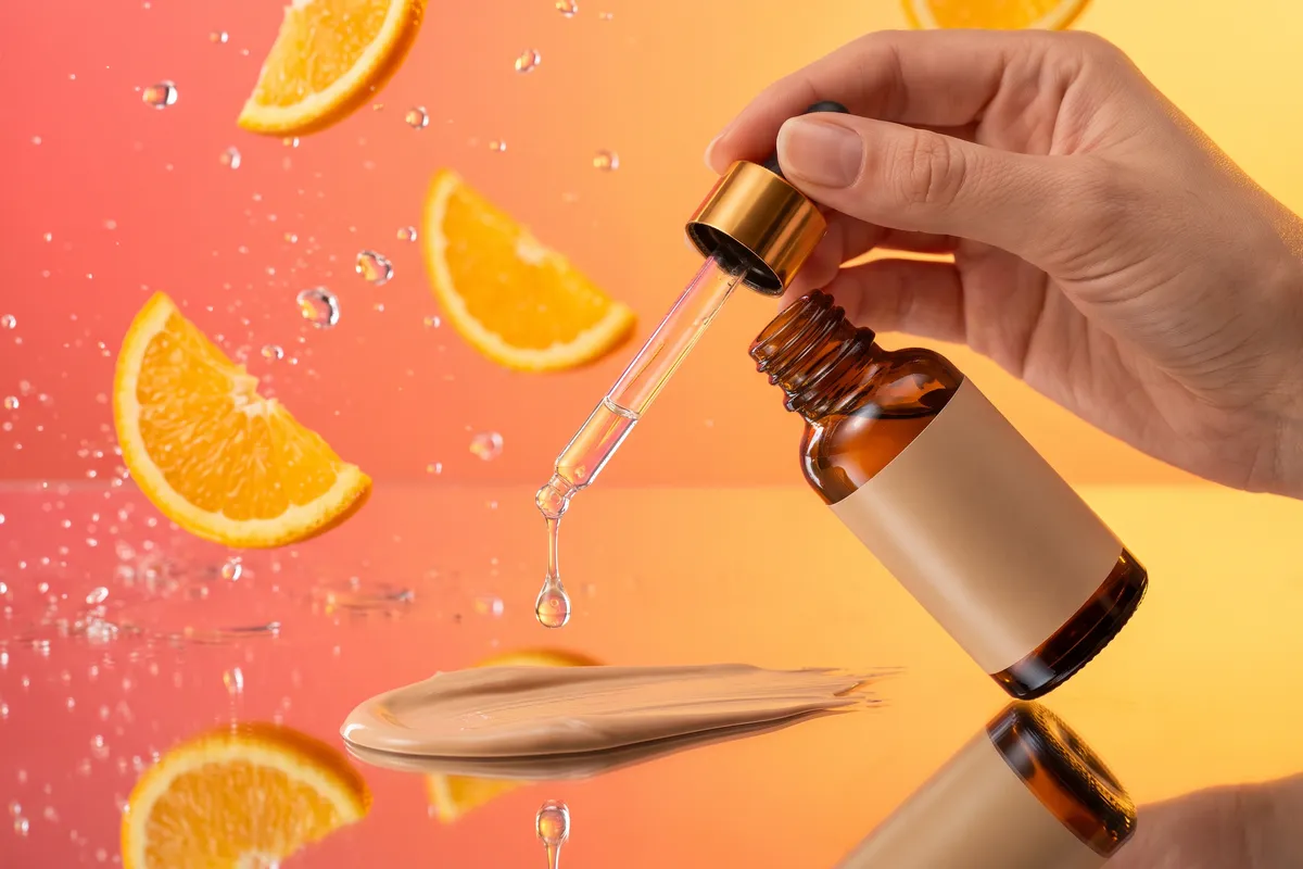 A hand holding an unbranded amber dropper bottle as a clear serum drop falls onto a swatch of tinted sunscreen, with orange slices, against a coral-to-gold backdrop