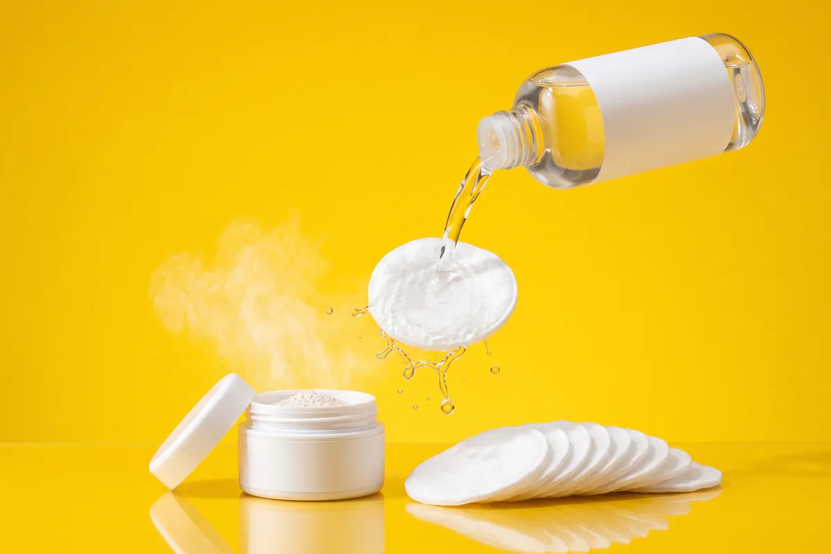 Unbranded glass toner bottle and an open powder exfoliant jar with a fan of cotton pads on a glossy surface against a sunny yellow backdrop