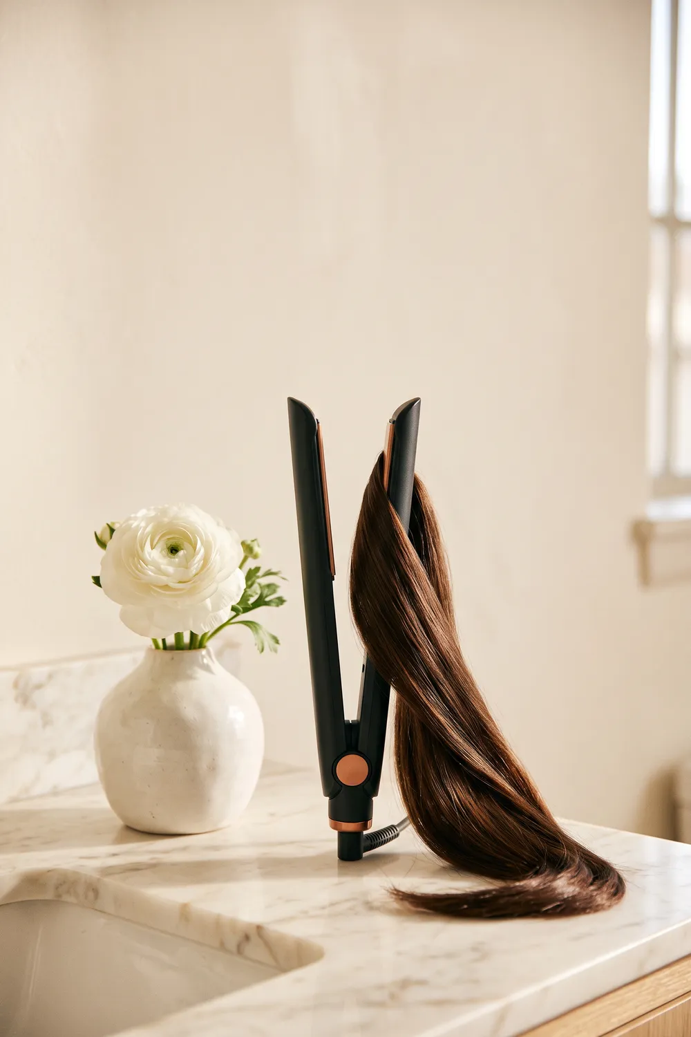 An unbranded nickel and copper air straightener with two slim arms, slightly open on cream linen on a pale marble vanity, beside a wide-tooth comb, a hair clip and a glossy straight strand of hair