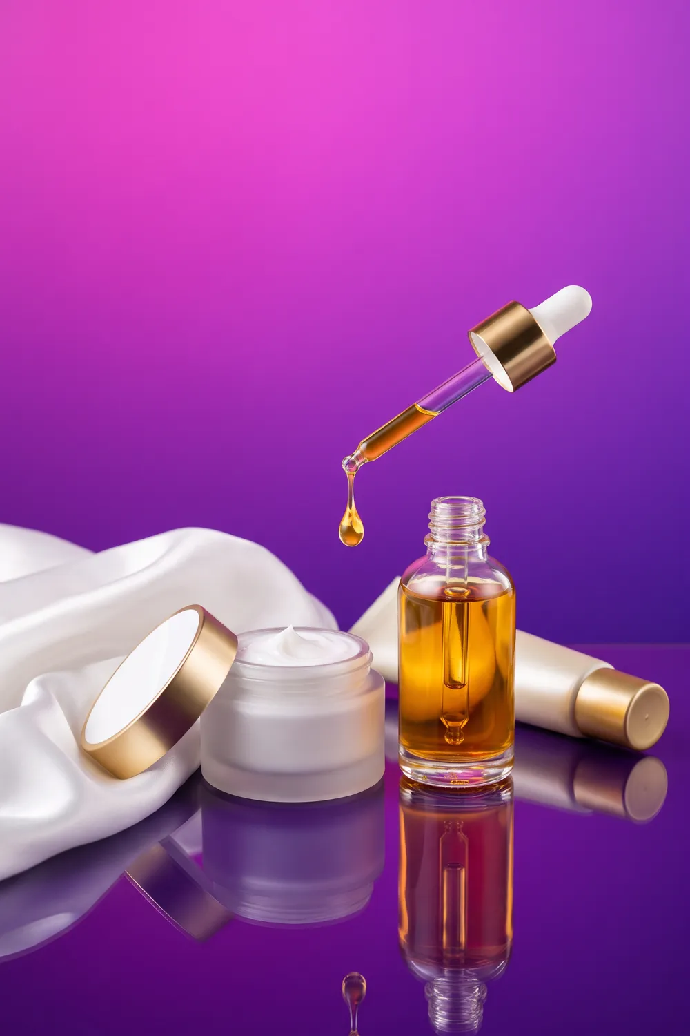 An unbranded sunscreen tube, a glass dropper bottle of amber serum and an open jar of glossy cream with a ribbon of white silk, on a deep violet to ultramarine backdrop