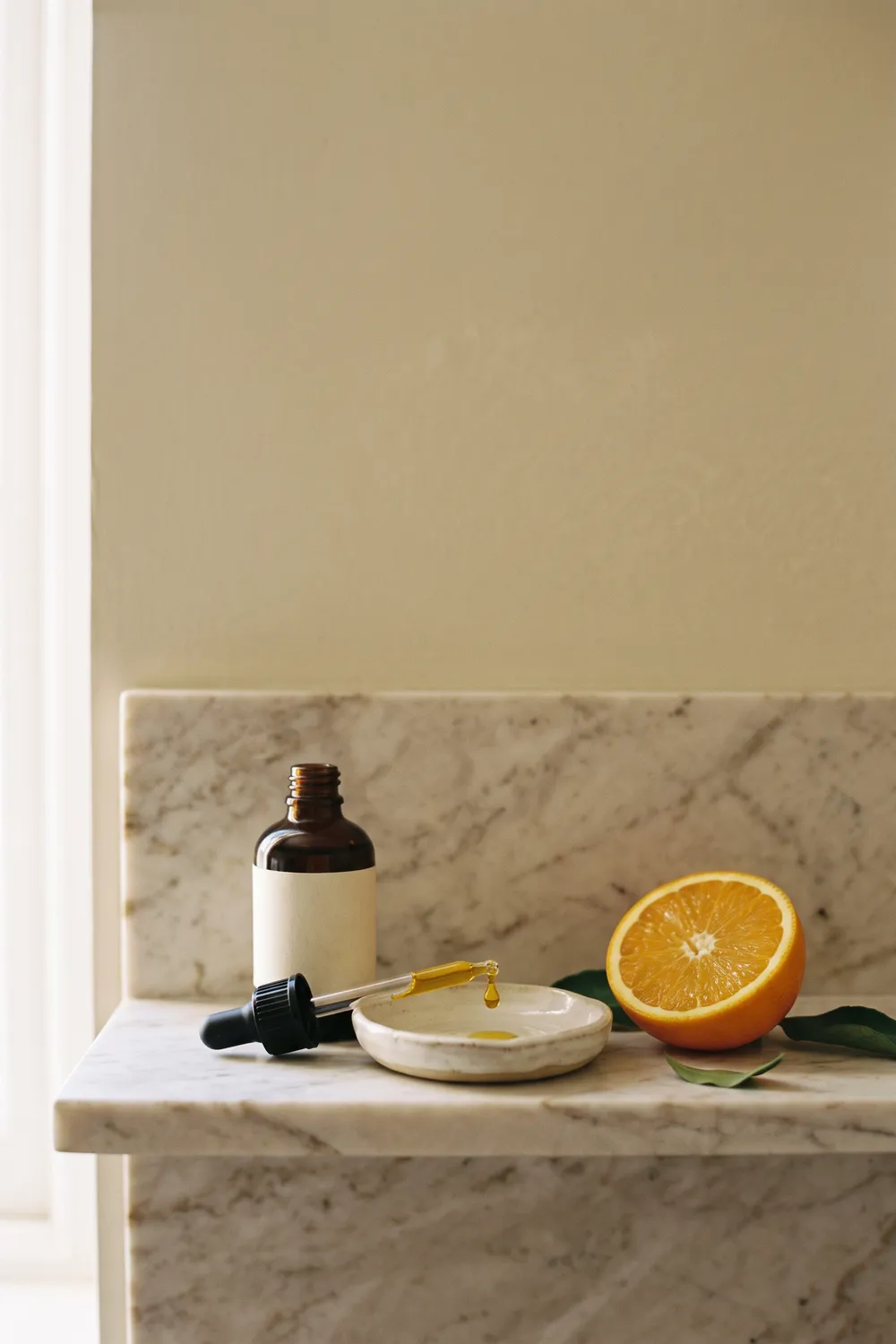 A small amber glass dropper bottle with a blank label beside a clear dropper bottle and a frosted moisturizer jar on a pale marble shelf, with sliced oranges, rosemary and drops of serum